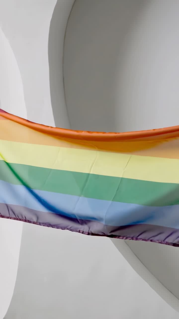 Video verticale: Brezza che solleva una bandiera arcobaleno che sventola e si raccoglie sulla corda, mostrando gli archi dell'atrio.