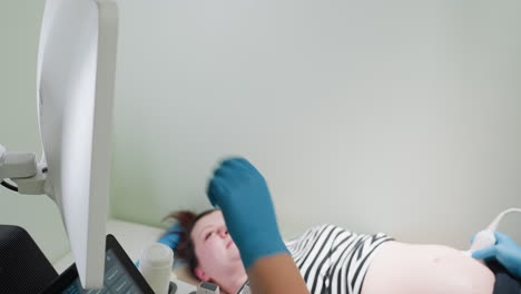 Female gynecologist wearing medical gloves turns monitor toward lying patient and gestures at screen while patient attentively observes examination results during interactive diagnostic consultation