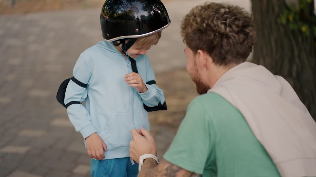 A little blond boy in a blue sweater does not want to wear a black protective helmet for riding a skateboard in a skate park, which his father with curly hair in a Green T-shirt put on him