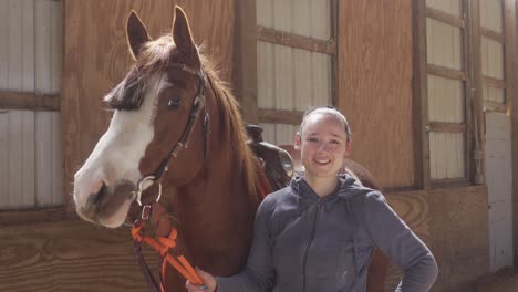 Beautiful-young-women-looking-into-the-camera-while-holding-a-pretty-white-faced-horse-in-western-bridal-and-saddle-with-a-blue-eye-inside-a-riding-arena-with-bright-warm-light-and-dust-4k-medium-shot