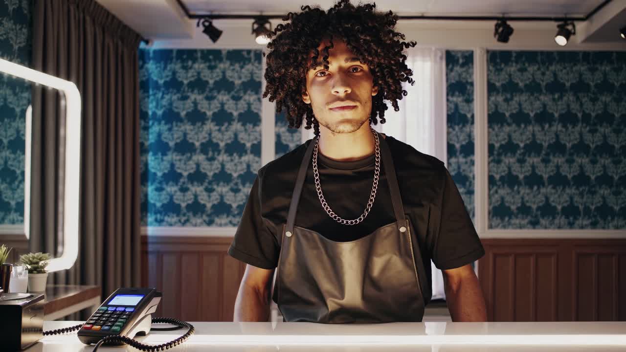 Professional hairdresser wearing black apron and silver necklace standing behind counter in vintage salon, waiting for potential clients with confident expression