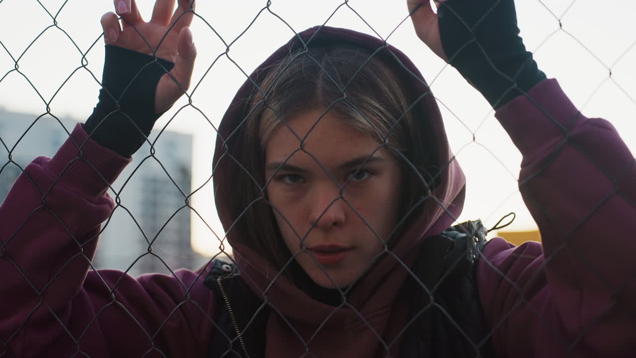 Fitness instructor in maroon hoodie and black vest rests hands on chain link fence while catching breath after intense urban lawn court workout with determination at sunset lighting city backdrop