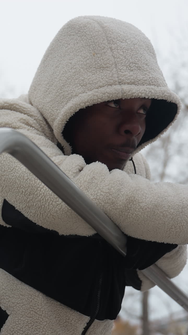 vista lateral de un hombre con suéter blanco descansando en barras de hierro, respirando pesadamente después de un intenso entrenamiento de invierno al aire libre, lleva guantes y una capucha, con una expresión enfocada en un parque nevado