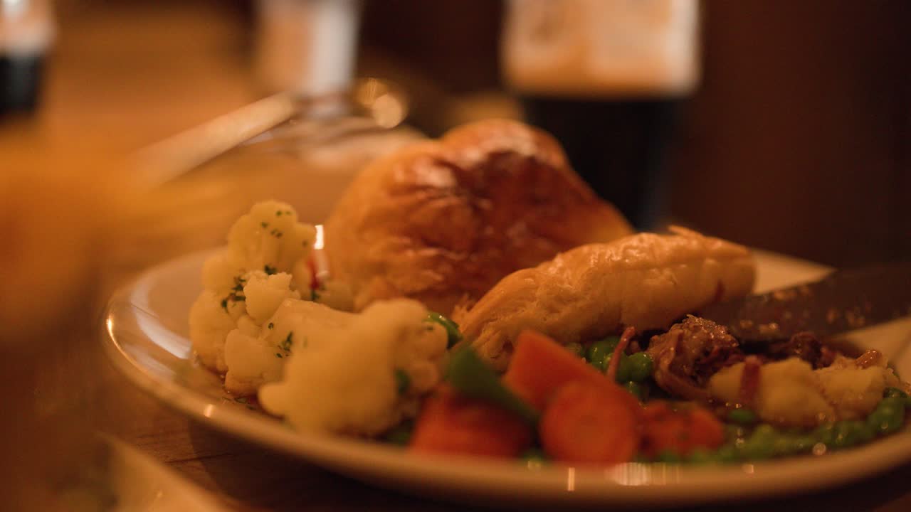 Hand slices steak pie on plate with vegetables, warm ambient lighting, shallow depth of field