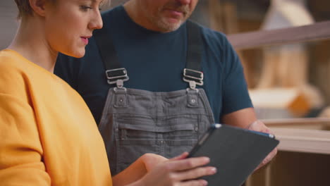 female apprentice learning from mature male carpenter with digital tablet in furniture workshop