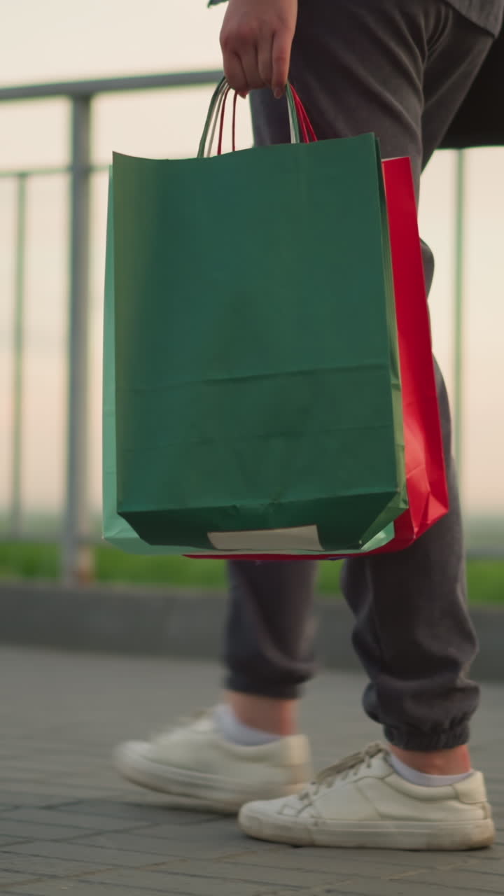 vista de la pierna de la dama en corredores grises caminando a lo largo del riel de hierro con bolsa de compras en la mano y bolso de mano negro, con fondo borroso suave con vegetación distante