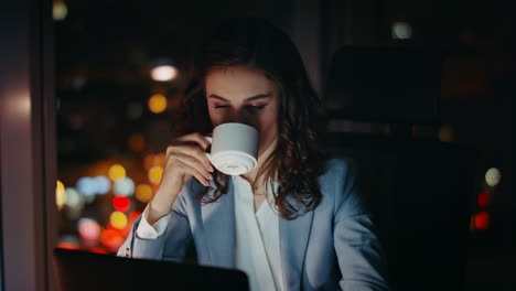 late worker drinking coffee on dark workplace looking laptop screen close up.