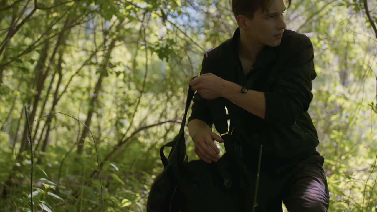 Tourist in dark outdoor clothing kneels on lush forest floor while turning head to right, preparing to change direction and open backpack, surrounded by thick green foliage and filtered sunlight