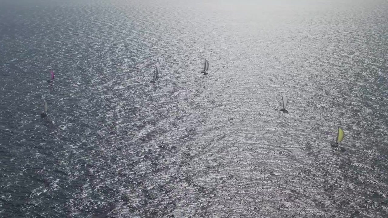 An elevated drone perspective capturing a group of colorful sailboats navigating open water during a competitive regatta under bright sunlight