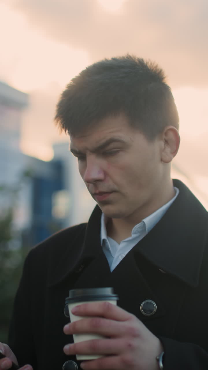 검은 코트를 입은 투자자가 야외에서 산책하고, 현대적인 건물, 주차된 자동차, 초록색, 그리고 걸어가는 사람들이 특징인 활기찬 도시 환경에서 커피를 마시면서 사려 깊게 드폰을 탐색하고 있습니다.