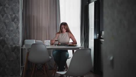 hungry woman wearing sleeveless top sitting at dining table eating sushi with food in her mouth, adjusting herself while crossing legs, enjoying meal alone in cozy modern kitchen