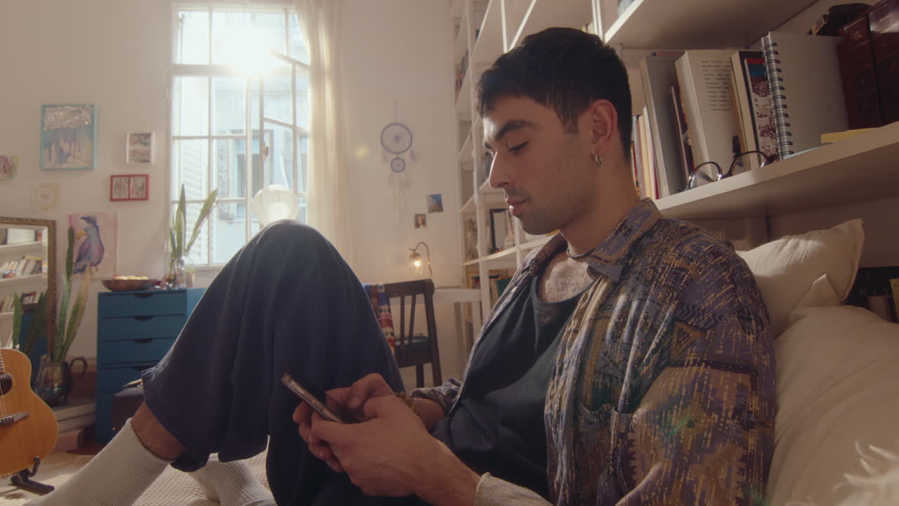 Young Man Sitting on Bed and Texting on Smartphone at Home