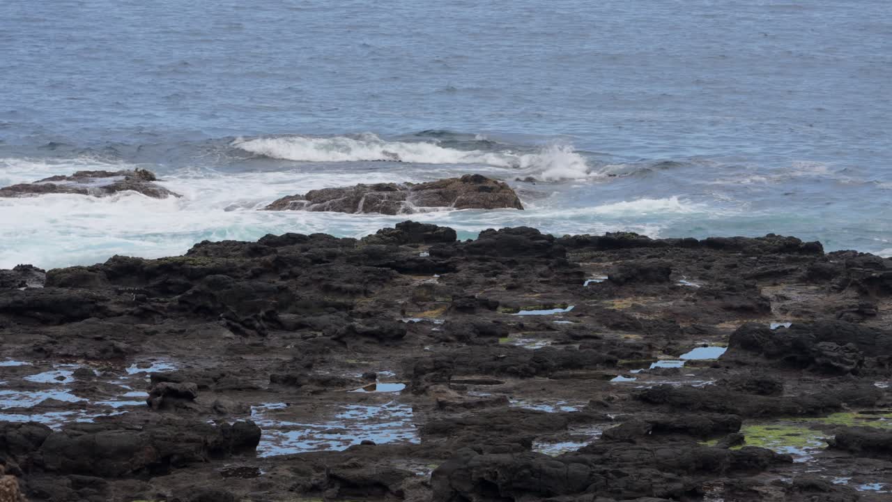 Ocean waves crash dramatically over a rugged rocky shoreline, creating a mesmerizing display of whitewater and splashes under the open sky.