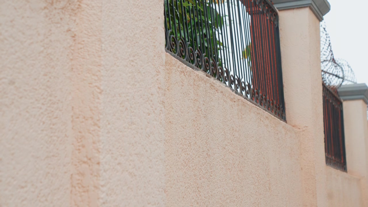 Close-up de parede de estuque com cerca de ferro mostrando exterior texturizado rosa pálido, grade decorativa de ferro forjado montada em coluna perimetral, vislumbres de folhagem verde através da treliça, luz do dia suave e nublada
