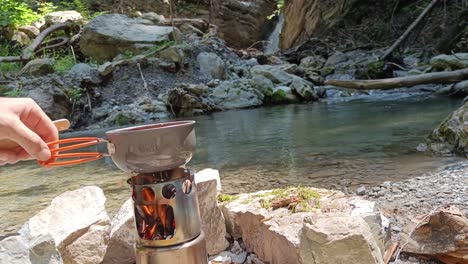 guy put pot with soup on hobo cooker and stir it with waterfall and river in background