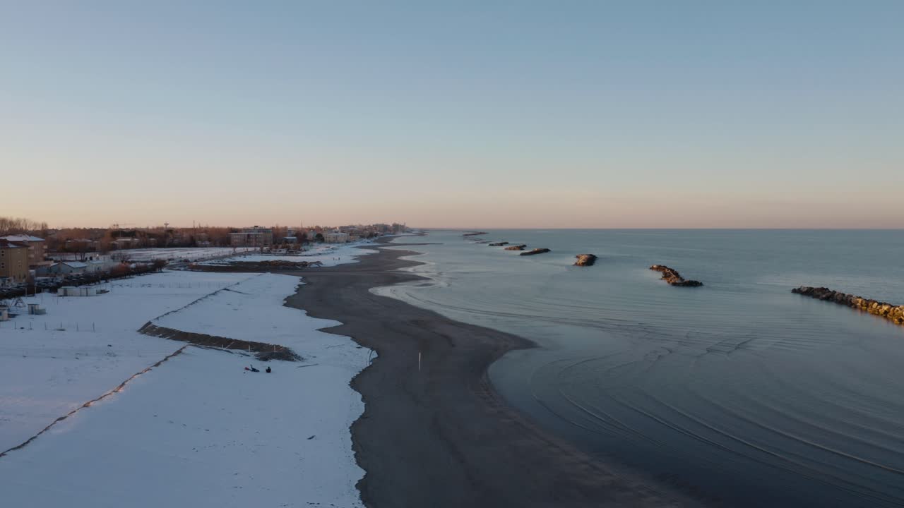 Bredt luftfoto af Riminis vinterkyst med sneklædt strand, mørk kystlinje, roligt hav og bølgebrydere under en blød pastel aftenhimmel