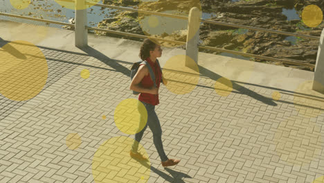 animation of yellow spots over biracial woman walking on promenade