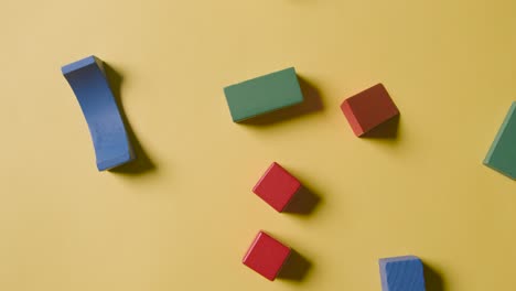 Overhead-Shot-Of-Colourful-Wooden-Building-Blocks-Being-Thrown-Onto-Yellow-Studio-Background