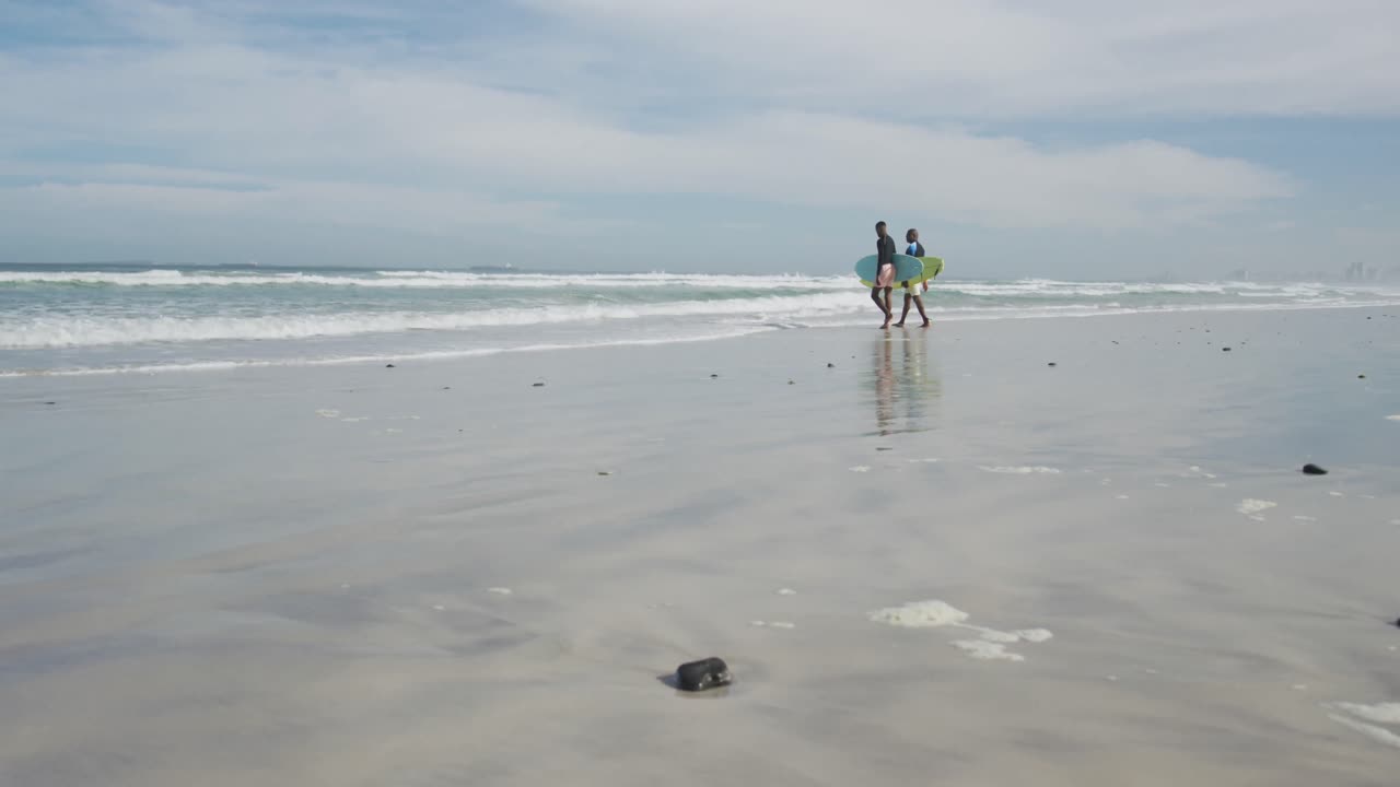 pai afro-americano e filho adolescente andando em uma praia segurando pranchas de surf e falando