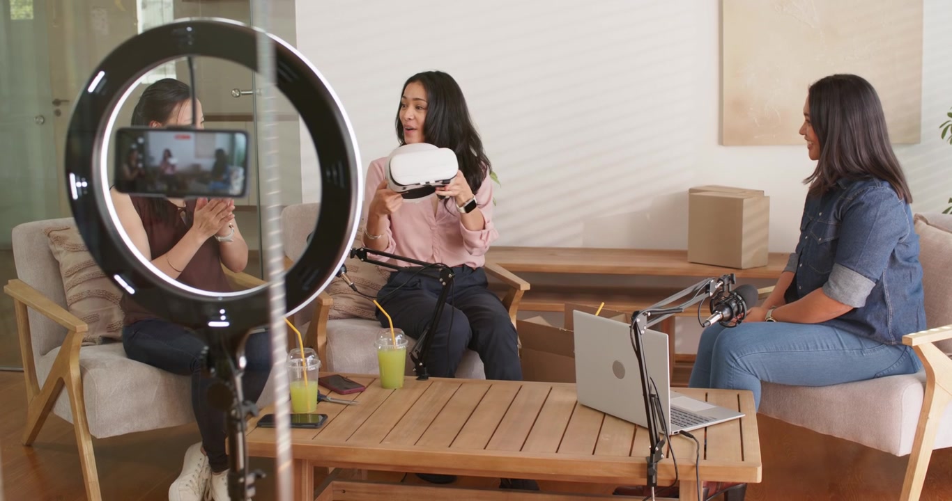 Presentadoras diversas en un estudio, la mujer asiática del centro abriendo una caja y mostrando un casco de realidad virtual a la cámara.