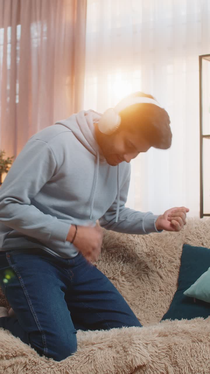 Young man dancing on couch with wireless headphones playing air guitar and enjoying disco music