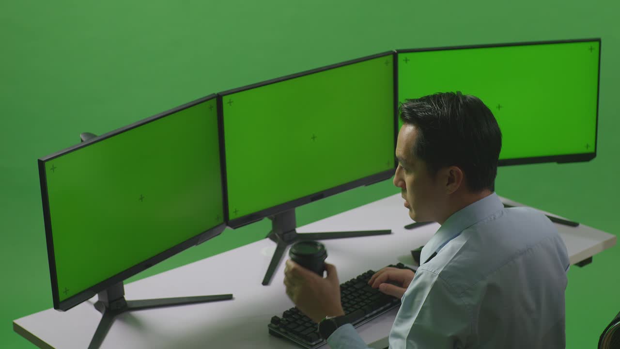 vista lateral de un hombre asiático bebiendo café y trabajando con un simulacro de múltiples monitores de computadora en un estudio de pantalla verde