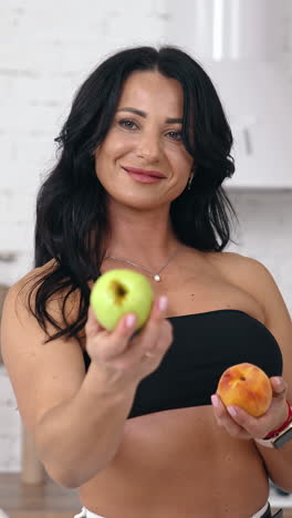 attractive female proposes fresh apples indoors. portrait of a woman with bare shoulders and belly holding juicy fruit and showing on camera in the kitchen. vertical video