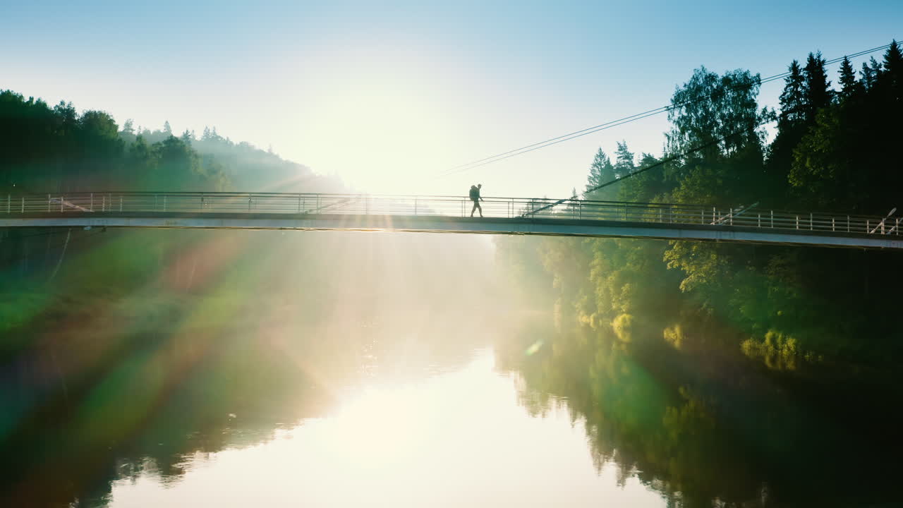 Solo backpacker crosses bridge on sunny morning, tracking drone shot