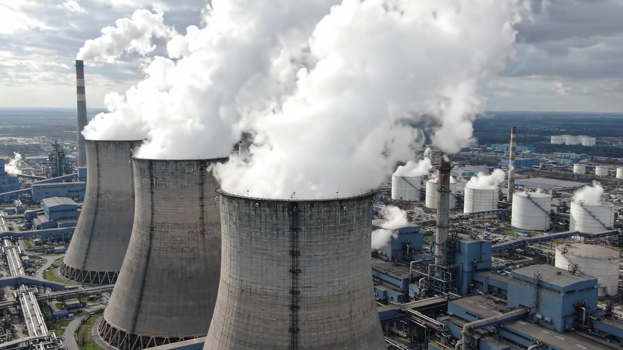 Vista aerea di una centrale elettrica industriale con torri di raffreddamento che emettono vapore e nuvole contro un cielo blu, mostrando la moderna tecnologia di produzione energetica