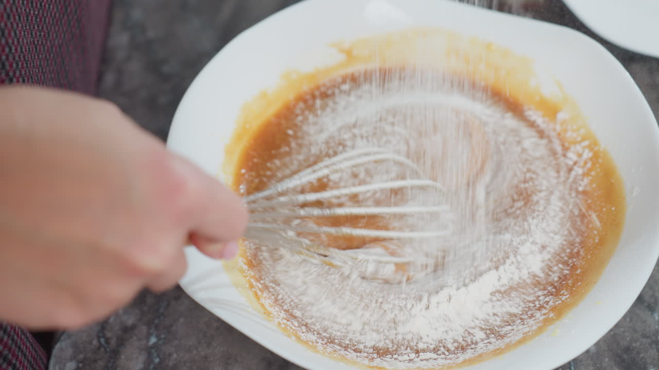 vista de arriba hacia abajo de una persona mezclando sopa espesa en un cuenco blanco mientras se tamiza la harina de maíz blanca fina, el batidor crea un efecto de mezcla suave, los ingredientes se combinan para una textura perfecta en la cocina casera
