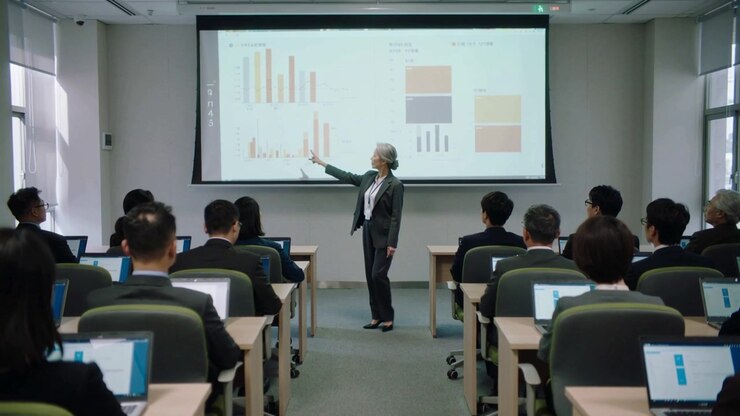 Slide advancing as trainer in suit walking front, pointing at charts showing data with laptops