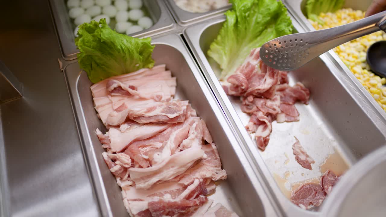 Hand uses tongs to pick raw sliced meat from buffet tray under bright kitchen lighting