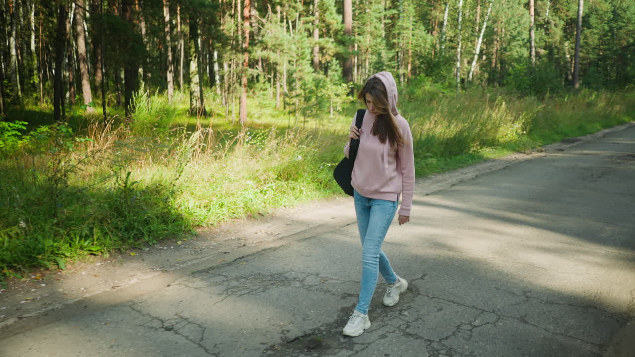 Beautiful woman in pink hoodie walking along quiet paved forest road, holding black bag strap with one hand, wearing jeans and sneakers, surrounded by lush green grass and tall trees under clear sky
