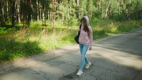 beautiful woman in pink hoodie walking along quiet paved forest road, holding black bag strap with one hand, wearing jeans and sneakers, surrounded by lush green grass and tall trees under clear sky