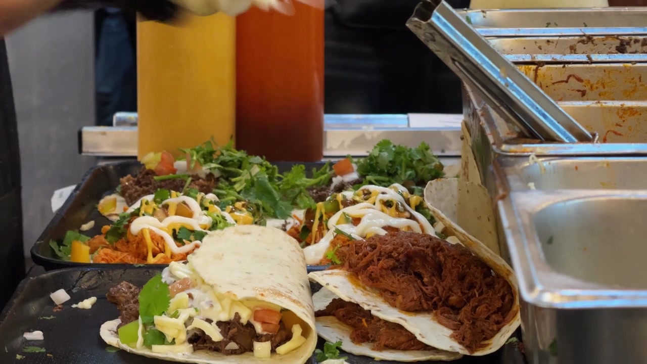 Nærbilledeoptagelse af en madbodssælger, der tilbereder flere friske tacos og wraps på en varm stegeplade. Tortillas fyldes med kød, ost, grøntsager, krydderurter og saucer under travl street food-tilberedning
