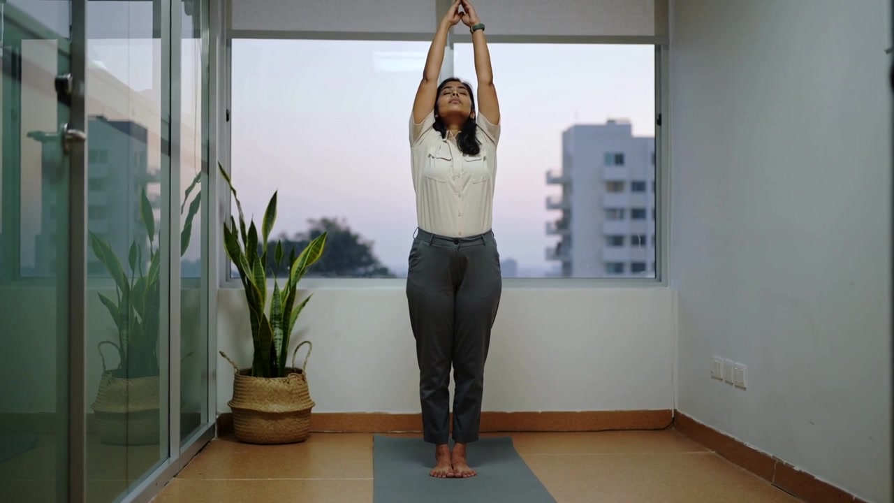 Mulher inspirando, de camisa branca e calças cinzas, levantando, unindo, abrindo e abaixando os braços no tapete do apartamento para se alongar.