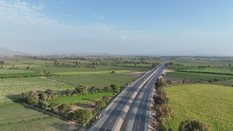 Toma-Aérea-Extremadamente-Larga-De-Vehículos-Que-Cruzan-La-Carretera-Panamericana-Norte-De-Cuatro-Carriles-Rodeada-De-Montañas-En-Perú-A-Través-De-Campos-Agrícolas-En-Ambas-Esquinas