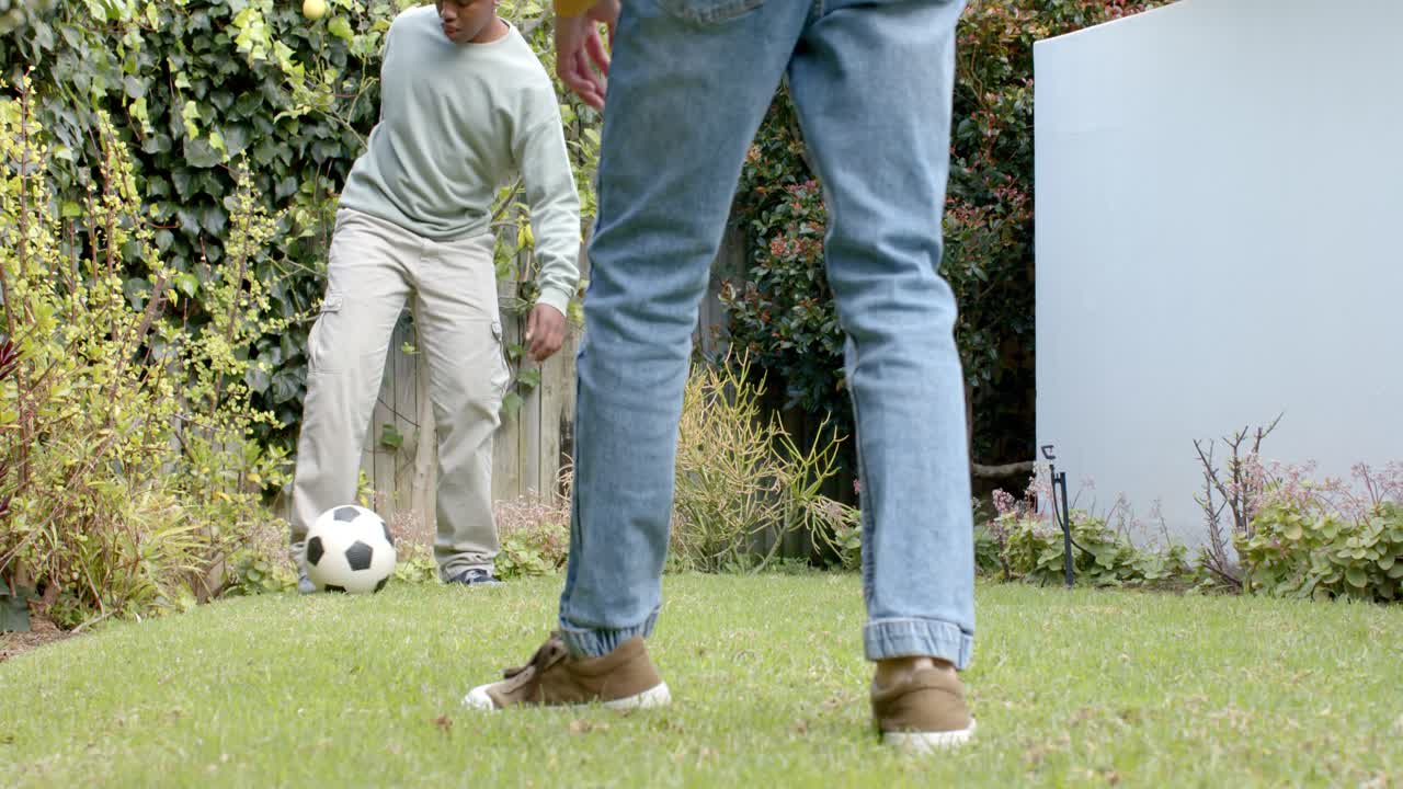 Diverse male friends nudging soccer ball to each other in backyard by ivy fence, practicing passes