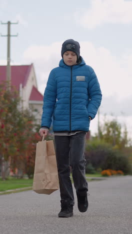 niño en chaqueta azul paseando por la calle residencial con mirada reflexiva, llevando una bolsa de papel, el fondo borroso presenta casas, árboles y vegetación