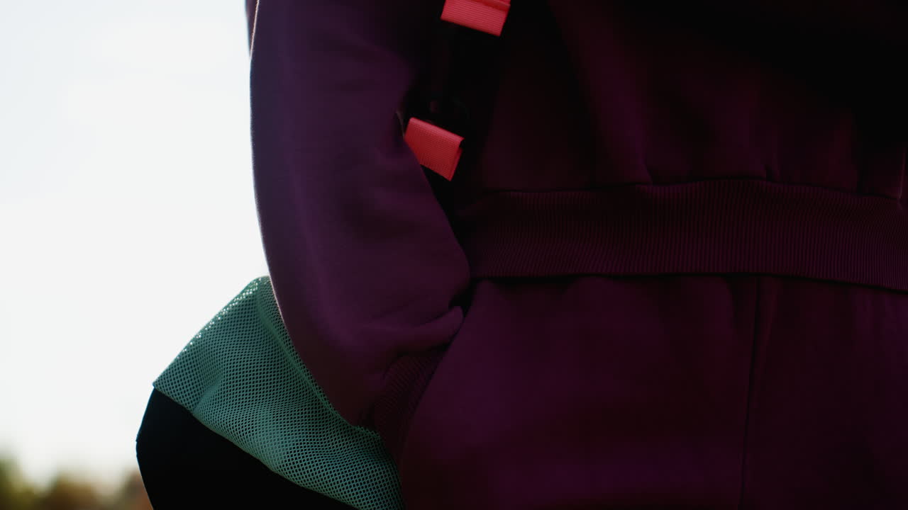 Elegant focused lady walking on gray tiled pavement under bright sunny sky casting soft shadows with building facade and autumn foliage backdrop capturing poised stride confidence in casual hoodie