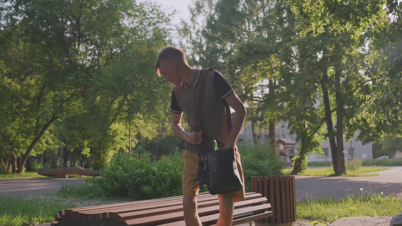 Un hombre caucásico examina sus pertenencias con concentración sobre un banco de madera. Busca meticulosamente en su bolsa mientras está sentado al aire libre bajo la luz del sol filtrada, mostrando un gran interés.