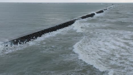 aerial establishing view of port of liepaja concrete pier , autumn storm, big waves splashing, overcast day, wide drone shot, orbit