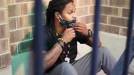 african american man with dreadlocks sitting on ground then pulls covid-19 mask out and putting in on, then waving with his phone, and getting up to leave