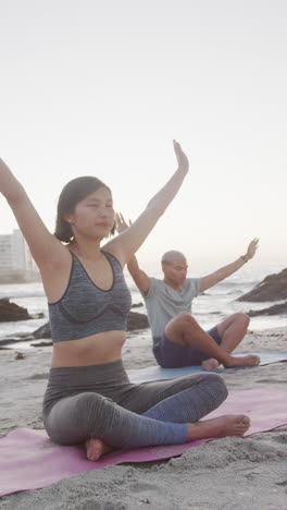 vertical video of happy biracial couple doing yoga and meditating at beach, in slow motion