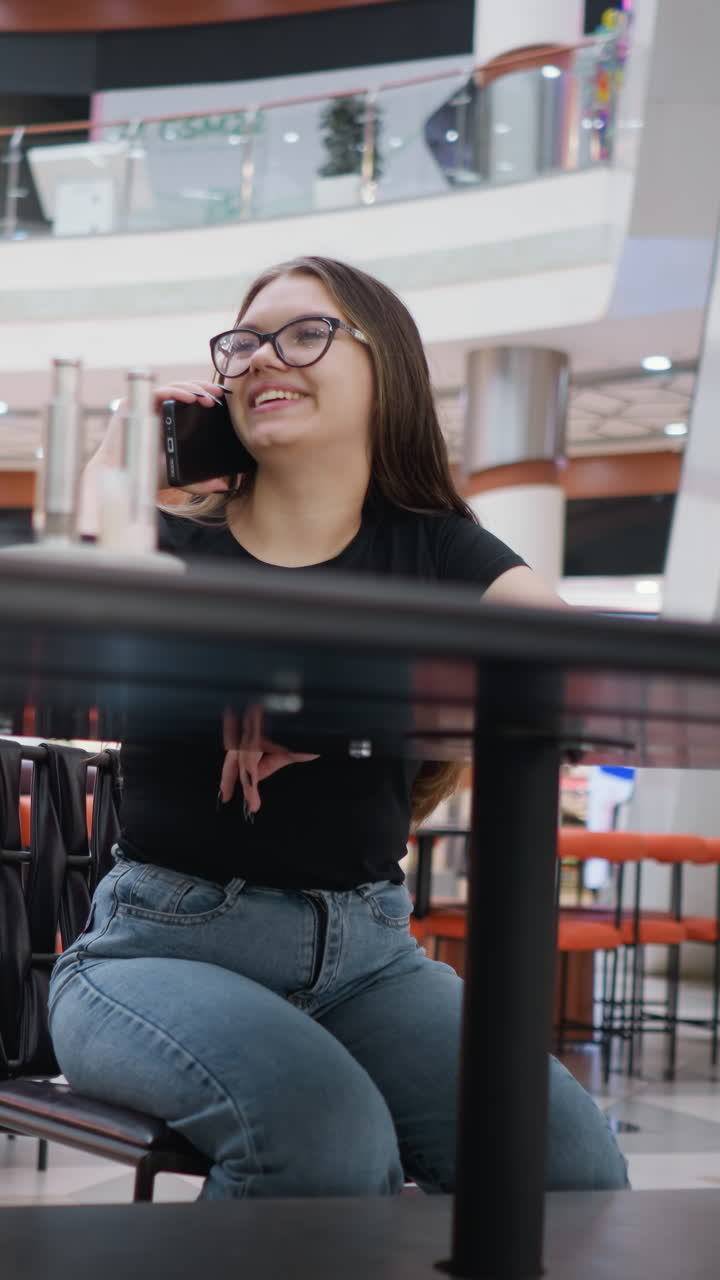 primer plano de sillas dispuestas con una dama con gafas involucrada en una llamada telefónica, disfrutando de la conversación en un centro comercial bellamente iluminado con inscripciones brillantes