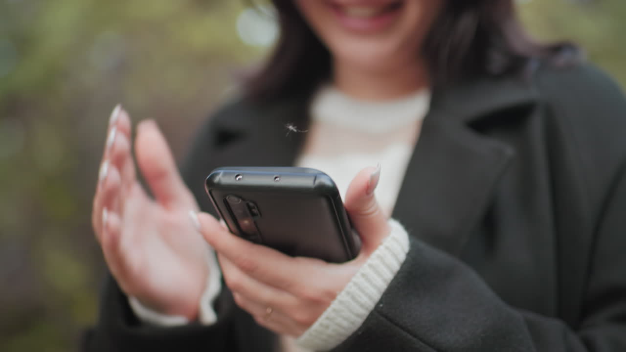 젊은 여성이 휴대폰 화면을 스와이프하며 웃고 있어요. 깔끔하게 관리된 손톱을 가진 손이 야외에서 휴대폰을 들고 있으며, 부드러운 보케 배경에 아늑한 코트 소매가 살짝 보이고, 현대적인 소통의 순간을 담은 낮은 앵글의 뷰입니다.