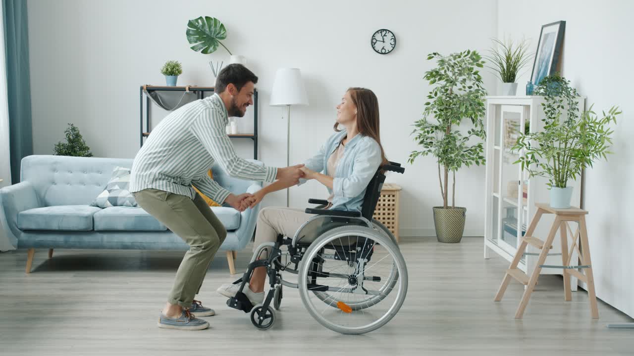 Slow motion of young disabled woman in wheelchair and joyful husband dancing having fun at home