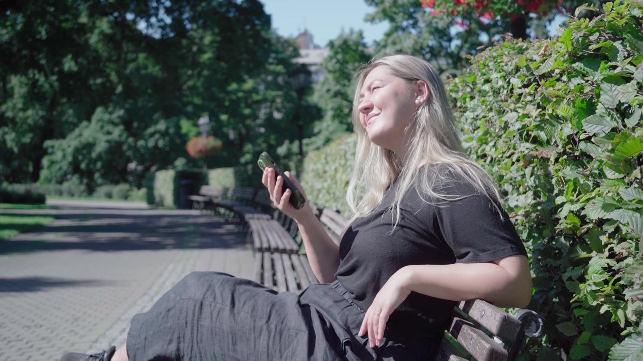 Smilende besøgende ligger tilbagelænet på en solrig parkbænk og tjekker rejseplaner på sin telefon midt mellem grønne hække og træer