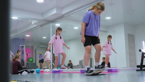 kids engage in balance and strength exercises in a gym, featuring a child in purple struggling to maintain balance, another seated, and one standing on one leg, large mirror reflecting the scene