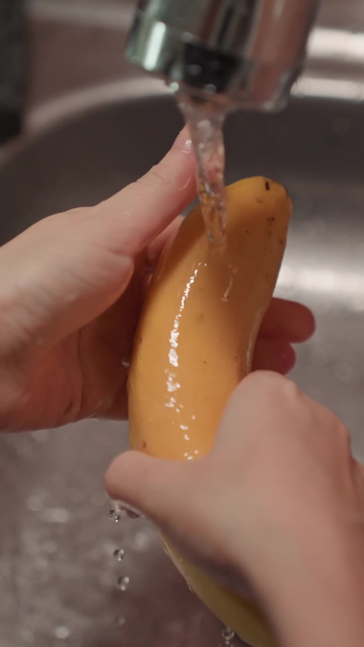 Primer plano de manos lavando un plátano amarillo bajo el agua corriente. La persona gira la fruta para limpiar todos los lados con líquido transparente. Fregadero de acero inoxidable y encimera oscura.